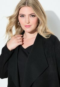 Schwarze Kunstwildlederjacke mit breitem Revers, kombiniert mit einem schwarzen V-Ausschnitt-Top. Das Model hat wellige blonde Haare und silberne Creolen.