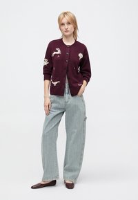 Burgundy knit cardigan with embroidered animal designs, button front, cropped sleeves, paired with light striped wide-leg jeans and flat shoes.