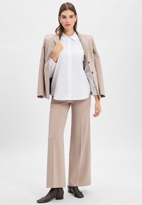 Vrouw in een beige blazer over de schouders, witte blouse met knopen, wijde beige broek en donkerbruine enkellaarzen op een effen achtergrond.