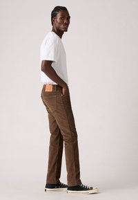 Pantaloni in velluto a coste marroni con vestibilità dritta, dotati di una toppa in pelle sul retro e sneaker nere. Abbinati a una t-shirt bianca semplice.