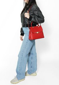 Borsa a tracolla in pelle rossa con finiture dorate, design strutturato, manici superiori e tracolla staccabile, abbinata a jeans blu casual e sneaker.