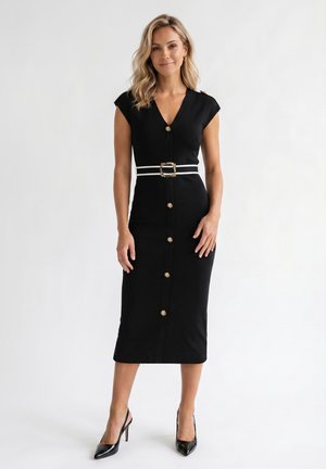Femme debout portant une robe noire ajustée avec des boutons dorés, une ceinture blanche avec une boucle dorée et des chaussures à talons noirs sur un fond blanc.