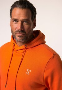 Hoodie in cotone arancione con cappuccio e cordini, presenta un piccolo logo bianco sul petto. Il tessuto sembra liscio ed è di un colore uniforme.
