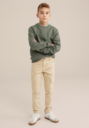 Felpa verde texturizzata, pantaloni cargo beige con tasche laterali e sneakers bianche con accenti beige. Design semplice e funzionale.