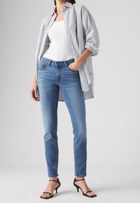 Kvinna i ljusblå slim fit-jeans, vit linne, överdimensionerad randig skjorta med knappar, och svarta öppna klackskor står med ena handen i fickan.