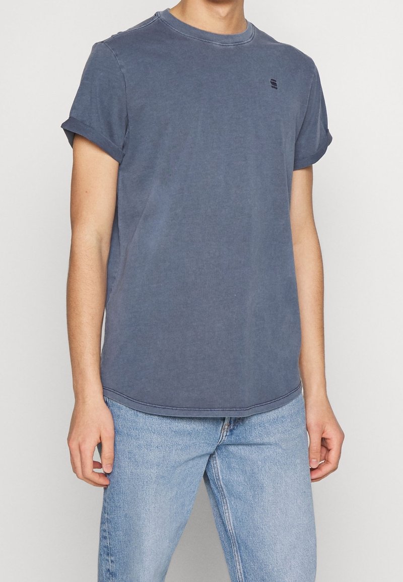 T-shirt en coton bleu avec des manches retroussées, col rond et un détail de logo subtil. Le tissu a une texture légèrement délavée, à assortir avec un jean en denim clair.