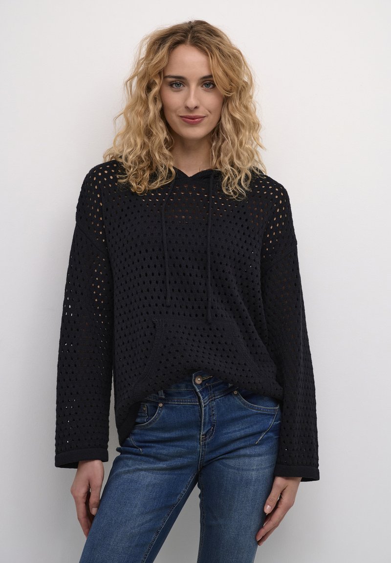 Melnā adītā kapuce ar brīvu piegriezumu, teksturētu acu rakstu, priekšējo kabatu un regulējamu kapuci; kombinēta ar zilo džinsu biksēm.