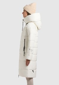 White puffer coat with a hood, quilted design, two side zippers, and black accents. Worn with a light beige knit beanie.