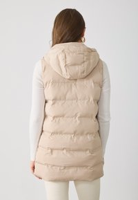 Gilet rembourré beige clair avec capuche, doté de sections matelassées horizontales et d'une texture lisse. Porté par-dessus un haut à manches longues.