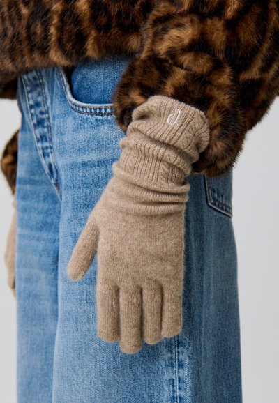 Guantes de punto beige con puño acanalado, mostrados en una mano. El conjunto acompañado incluye una chaqueta de piel de marrón y denim azul.