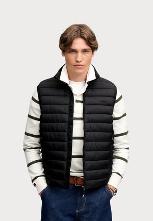 Junger Mann trägt eine schwarze gesteppte Weste über einem weiß-grün gestreiften Langarmshirt, blaue Jeans und einen braunen Gürtel, steht mit den Händen in den Taschen.