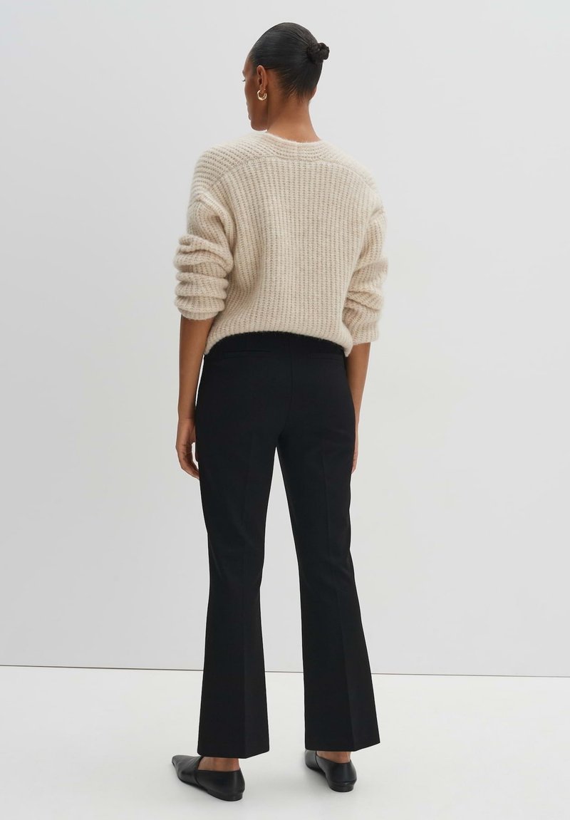 Beige gestrickter Pullover mit einem strukturierten Muster, kombiniert mit schwarzen maßgeschneiderten Hosen und schwarzen Ballerinas, von hinten betrachtet.