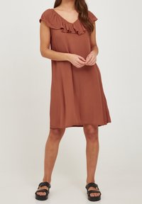 Roodbruin jurkje met V-hals en ruffles bij de halslijn, korte mouwen en een vloeiende snit, gecombineerd met zwarte sandalen voor contrast.