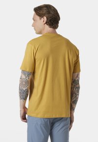 Geel katoenen T-shirt met korte mouwen, ronde halslijn en een effen achterkant. Zichtbare tatoeages op de arm. Gedragen met lichtblauwe broeken.
