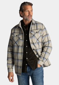 Beige geruite flanellen shirt-jacket met blauwe en zwarte lijnen, voorzien van twee borstzakken, zwarte knopen en een zachte textuur.