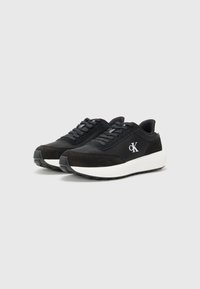 Zwarte sneakers met een gladde en suède textuur, voorzien van een witte tussenzool, grijze buitenzool en een logo aan de zijkant. Veters en een laag profiel ontwerp.