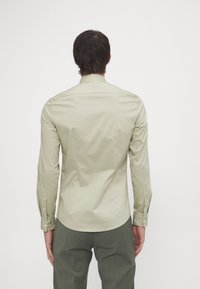 Uomo con capelli corti scuri, che indossa una camicia a maniche lunghe beige chiaro con bottoni e pantaloni verde oliva, mostrato di spalle.