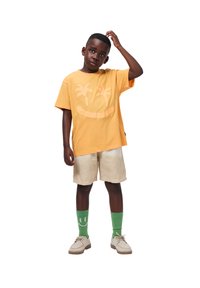 Narancssárga pamut póló pálmafával és mosolygós arccal díszítve, bézs rövidnadrággal és zöld mintás zoknikkal párosítva. A laza lábbeli teszi teljessé a megjelenést.