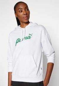 Sudadera blanca hecha de una tela suave, con un logo verde de "Puma" y un bolsillo tipo canguro. Incluye puños y dobladillo acanalados para un ajuste ceñido.