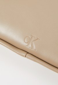 Borsa in pelle beige con texture liscia, caratterizzata da un logo "ck" impresso e bordi strutturati e arrotondati. I dettagli mettono in risalto il design minimalista.