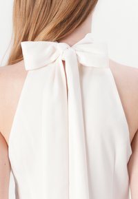 Gaiša krēmu halter stila blūze ar lielu kakla loku aizmugurē. Gluda, plūstoša auduma ar mīkstu tekstūru un atvērtu aizmuguri.