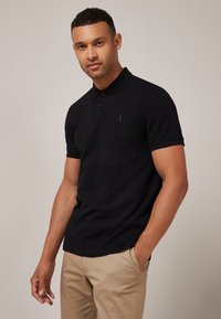 Sort polo t-shirt med korte ærmer, lavet af tekstureret stof. Har en krave og et diskret logo på brystet, kombineret med beige bukser.