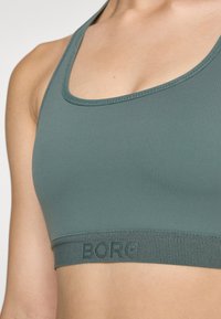Sujeción deportiva en tejido verde con una textura suave. Cuenta con un escote redondeado y una banda elástica gruesa con el texto "BORG".