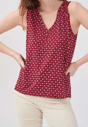 Haut rouge sans manches avec petit imprimé géométrique blanc, porté avec un pantalon beige clair, mains posées sur les hanches.