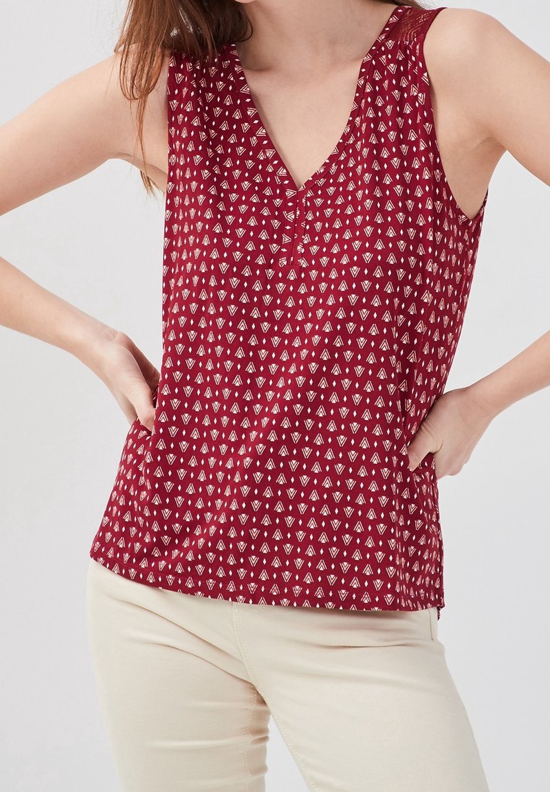 Haut rouge sans manches avec petit imprimé géométrique blanc, porté avec un pantalon beige clair, mains posées sur les hanches.