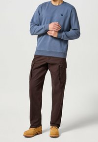 Ljusblå sweatshirt med rund hals och ribbade muddningar, tillsammans med mörkbruna cargobyxor med sidofickor och avslappnad passform. Tan-kängor.