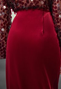 Vestido de terciopelo rojo con una silueta ajustada. Parte superior transparente adornada con lentejuelas circulares de diferentes tamaños. Detalle de cintura acentuado.