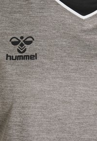 Grå sportsbluse lavet af tekstureret stof, med et sort "hummel" logo og en sort halsudskæring med hvid kant.