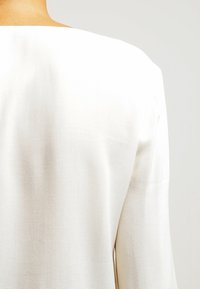 Weiße Bluse aus glattem, strukturiertem Stoff mit weitem Ausschnitt und langen Ärmeln. Das Design legt Wert auf Schlichtheit und Eleganz.