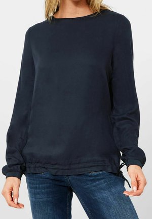 Femme portant un chemisier bleu marine à manches longues avec poignets et ourlet froncés, associé à un jean en denim bleu, vue des épaules aux hanches.