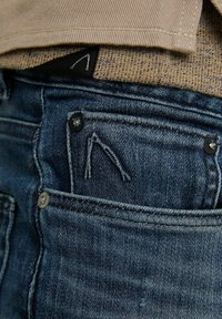 Jean en denim bleu foncé avec une poche avant ornée de surpiqûres foncées et de ferrures métalliques, partiellement cachée par une couche supérieure beige.