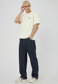 T-shirt en coton crème à manches courtes, associé à un pantalon en denim bleu foncé avec des poches latérales et des coutures jaunes contrastantes.