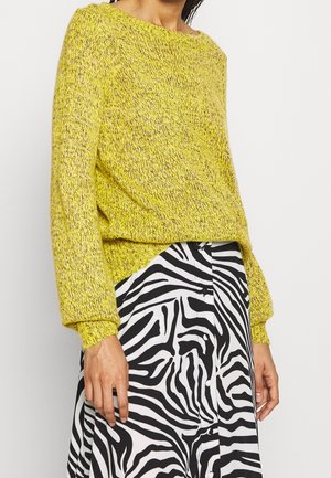 Personne portant un pull en maille jaune associé à une jupe boutonnée imprimé zèbre noir et blanc, debout devant un fond uni.