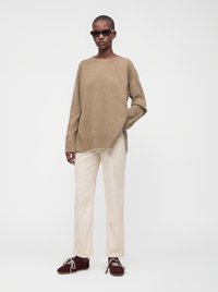 Femme portant un pull marron, un pantalon crème, des chaussures marron foncé et des lunettes de soleil, debout devant un fond blanc.