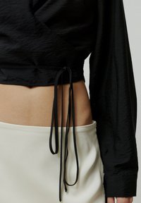 Schwarzes Cropped Top mit langen Ärmeln, aus strukturierter Stoffqualität, mit seitlichem Bindedetail und kontrastierendem weißen Taillenbund eines Rockes darunter.
