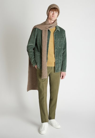 Chaqueta de pana verde sobre un suéter amarillo, pantalones oliva y zapatillas blancas; acompañada de una bufanda de punto marrón y un gorro.