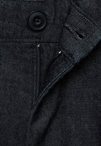 Close-up van zwart denim stof met een grote knoop, rits en een opvallend diagonaal weefpatroon. Stiksels zijn zichtbaar.