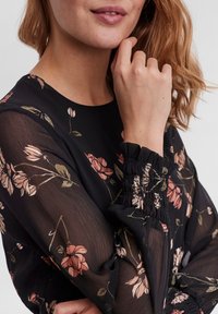 Blusa negra transparente con estampado floral en rosa y verde, que presenta largas mangas abullonadas con puños elásticos y un escote redondeado.