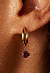 Gouden oorbel met een ronde, roze edelsteen hangend aan de ring, tegen een gestructureerde huid achtergrond.