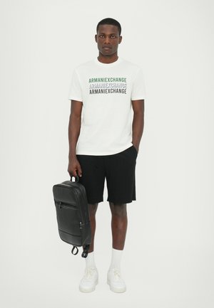T-shirt blanc avec le texte « ARMANIEXCHANGE » répété en bleu et vert, shorts noirs, baskets blanches et un sac à dos en cuir noir.