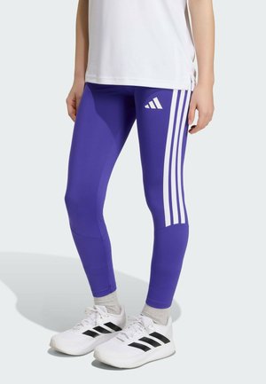 Persoon draagt paarse Adidas legging met witte strepen, grijze sokken, witte Adidas sneakers en een wit shirt, staand op een lichte achtergrond.