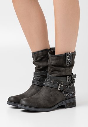 Harmaat mokkanahasta nilkkurit, joissa käännetyt varret, koristehihnat, niitit ja teksturoitu yksityiskohta. Tukevat pohjat matalalla korolla.