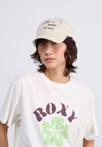 Beige caps med brodert tekst "MAKE IT MAKE SENSE." og en kremfarget T-skjorte med en mørk lilla "ROXY"-logo med et grønt blomsterdesign.
