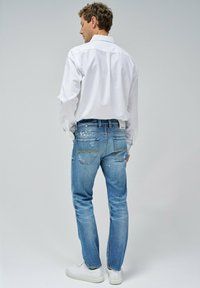 Jeans de mezclilla azul claro con un ajuste relajado, que presentan detalles de salpicaduras de pintura en las piernas y costuras amarillas en los bolsillos traseros. Combinados con una camisa blanca.
