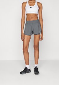 Pantalones cortos deportivos grises con una tela ligera y transpirable, con un logo de Nike en el dobladillo. Combinados con un sujetador deportivo blanco y zapatillas deportivas negras.