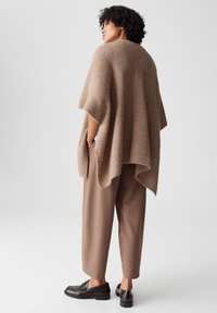Beige gebreide poncho met een geribbelde textuur, met een losse pasvorm en zijzakken, gecombineerd met een bruine broek en zwarte schoenen.